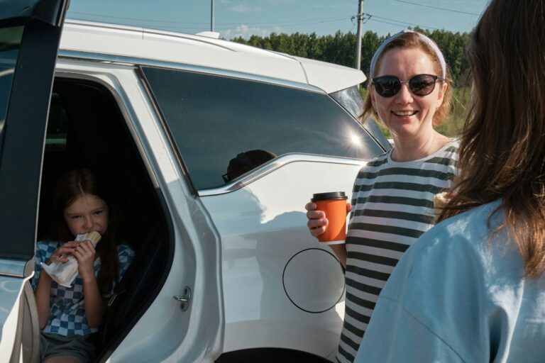 Mother standing beside a car holding a coffee while speaking with another adult, with a child sitting inside the vehicle in a busy family setting