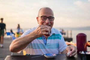 Older man sitting outdoors by the water smiling and holding a drink, appearing relaxed and at ease
