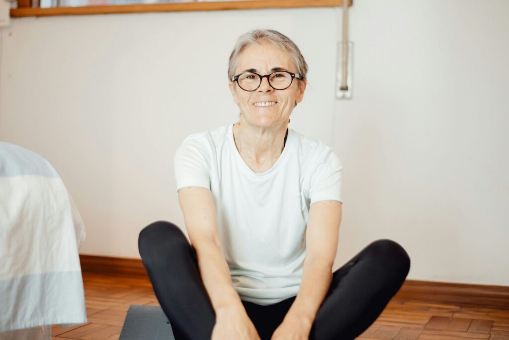 Older woman sitting on a yoga mat indoors, smiling calmly and appearing relaxed and at ease
