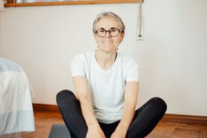 Older woman sitting on a yoga mat indoors, smiling calmly and appearing relaxed and at ease